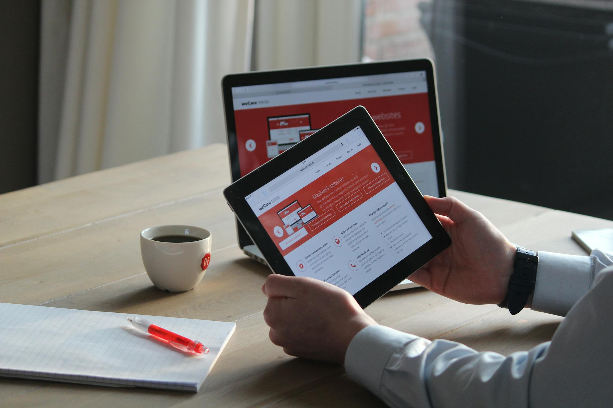 Business professional using a tablet and laptop with a hot drink, focusing on digital content review.